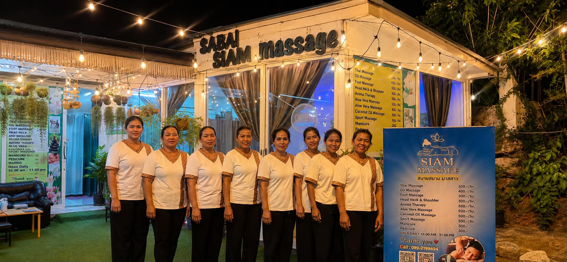 The eight therapists of Sabai Siam lined up in front of the lit salon at night, in matching white and Thai-patterned uniforms.